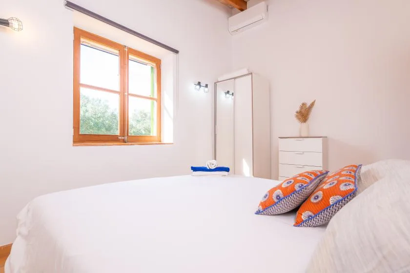 A bright bedroom with a large bed, colorful pillows, and a wooden-framed window.