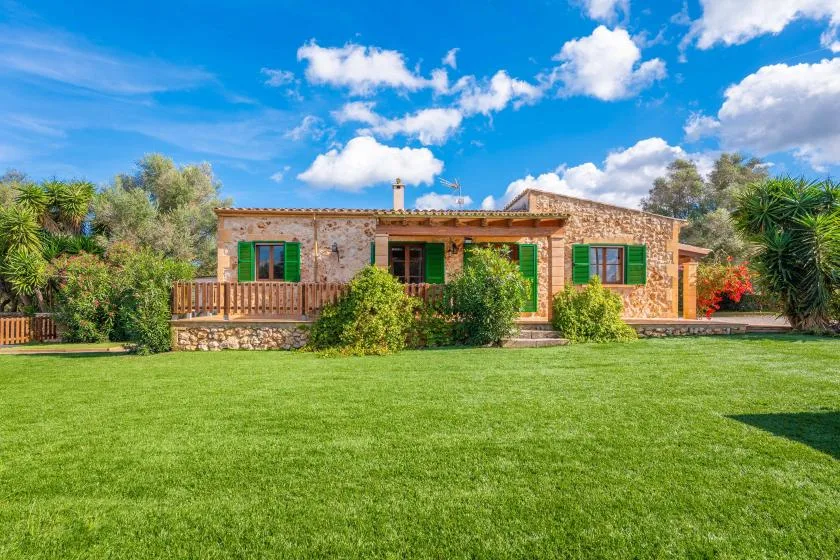 A stone house with green shutters faces a broad lawn and surrounding trees.