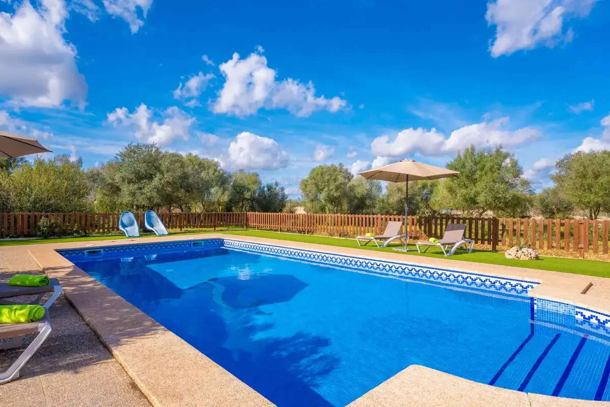 A swimming pool bordered by a wooden fence, with loungers and an umbrella.