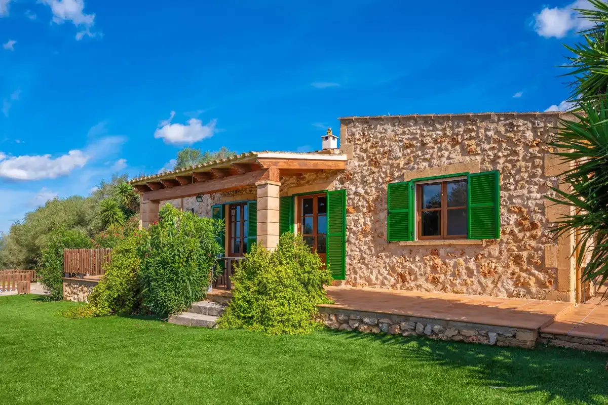 A stone house with green shutters stands beside a manicured lawn.