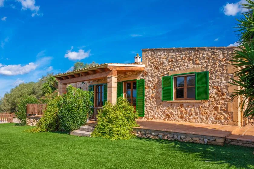 A stone house opens onto a green lawn, with green shutters and a shaded patio.