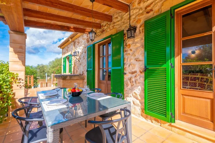 A covered outdoor dining area with a glass table beside stone walls and green shutters.