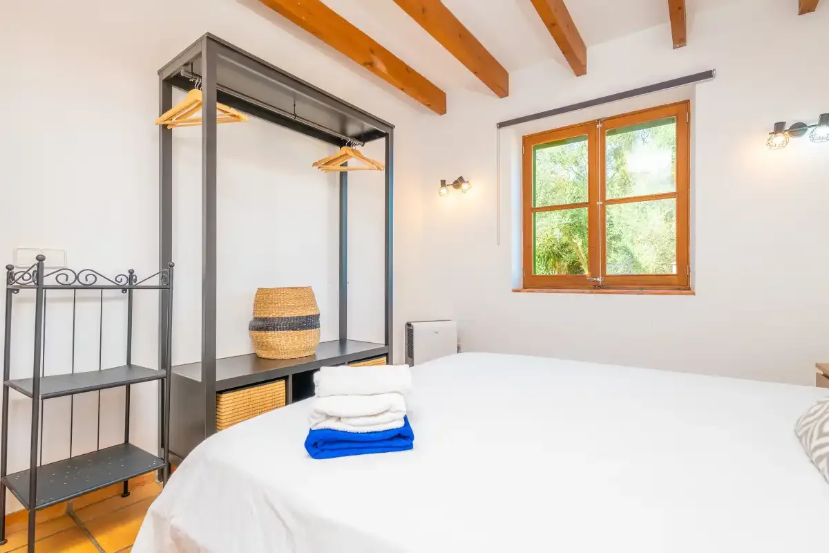 A bedroom with a neatly made bed, open clothes rack, and wooden-framed window.