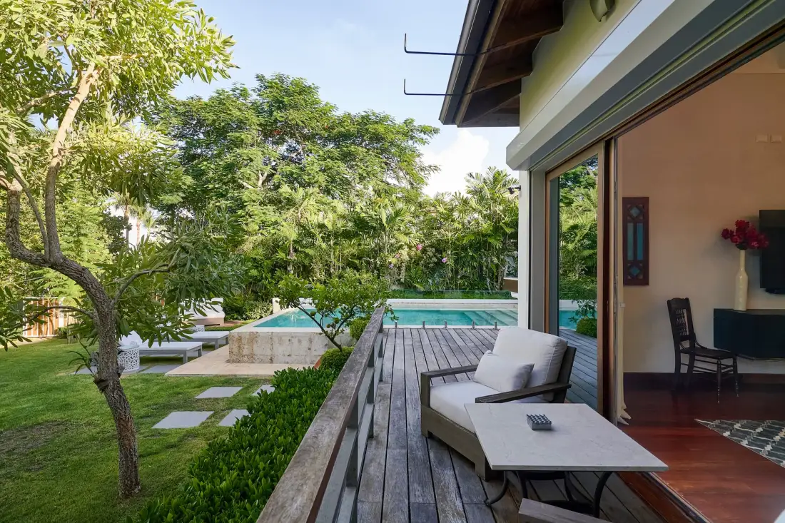 A patio overlooks a swimming pool and leafy garden beside a glass-walled room.
