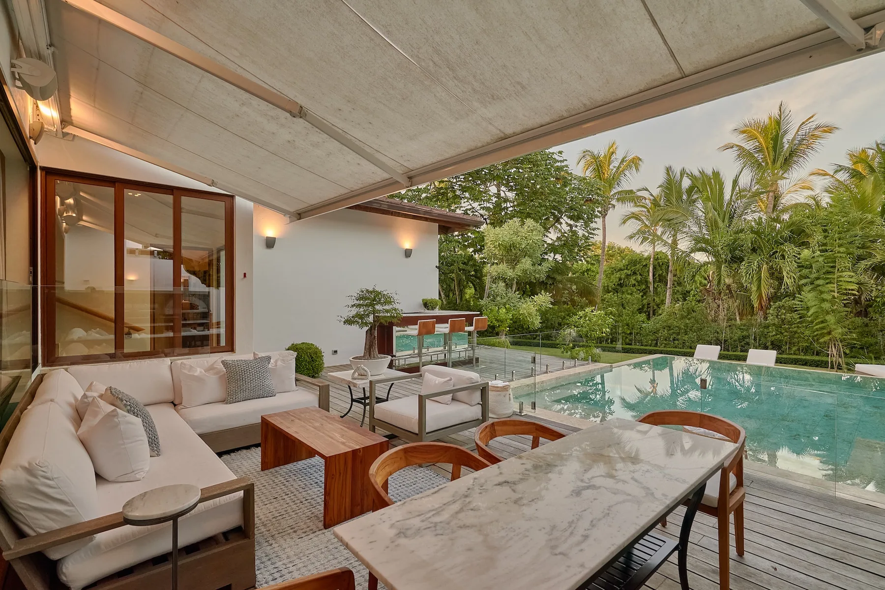 A covered patio with outdoor seating and a swimming pool surrounded by greenery.