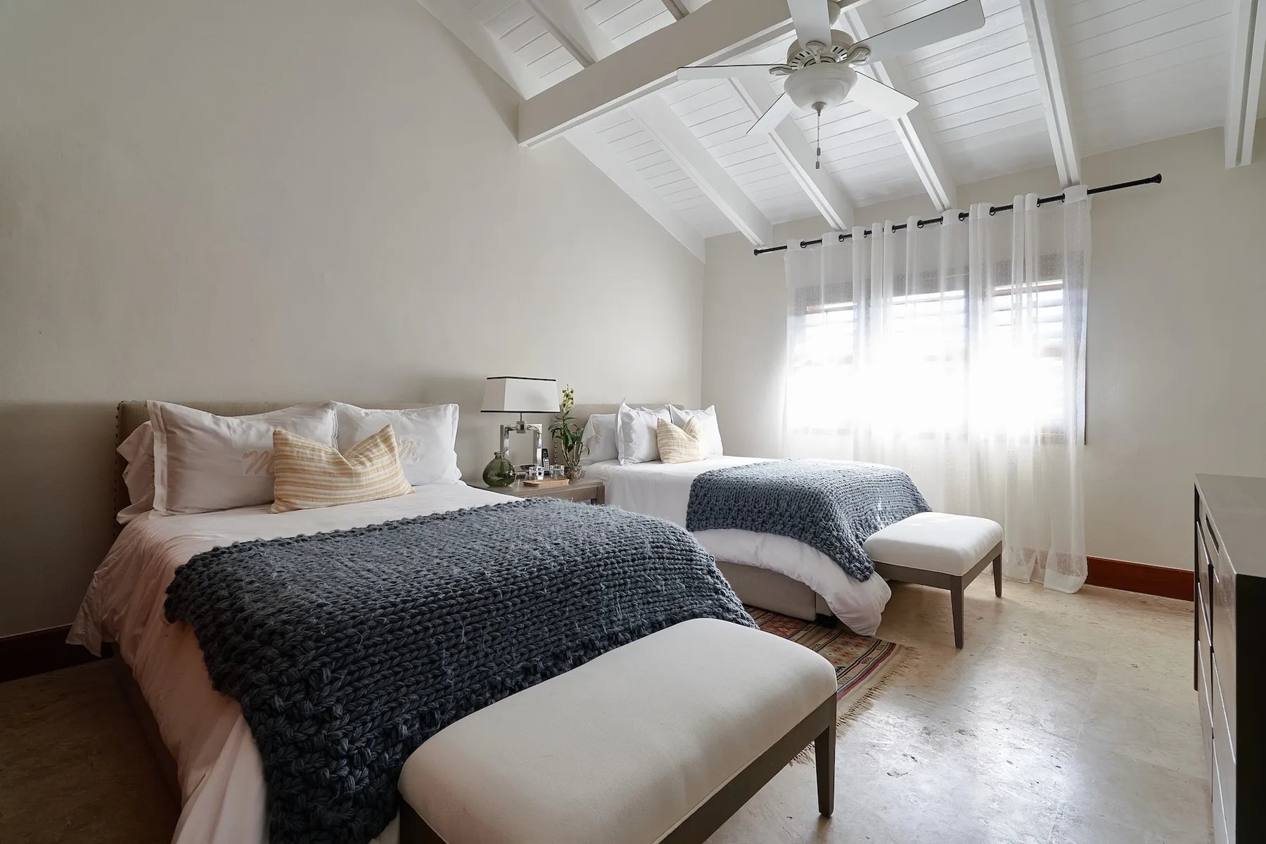 A bright bedroom with two beds, white curtains, and a vaulted ceiling.