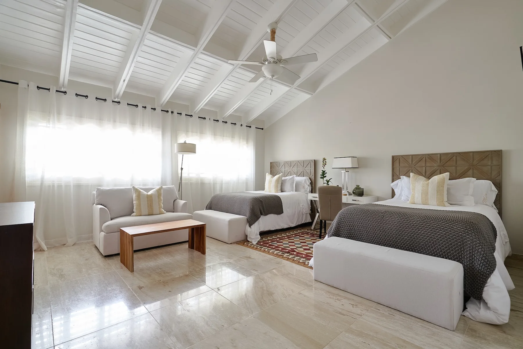 A bright bedroom with two beds, a seating area, and a white vaulted ceiling.
