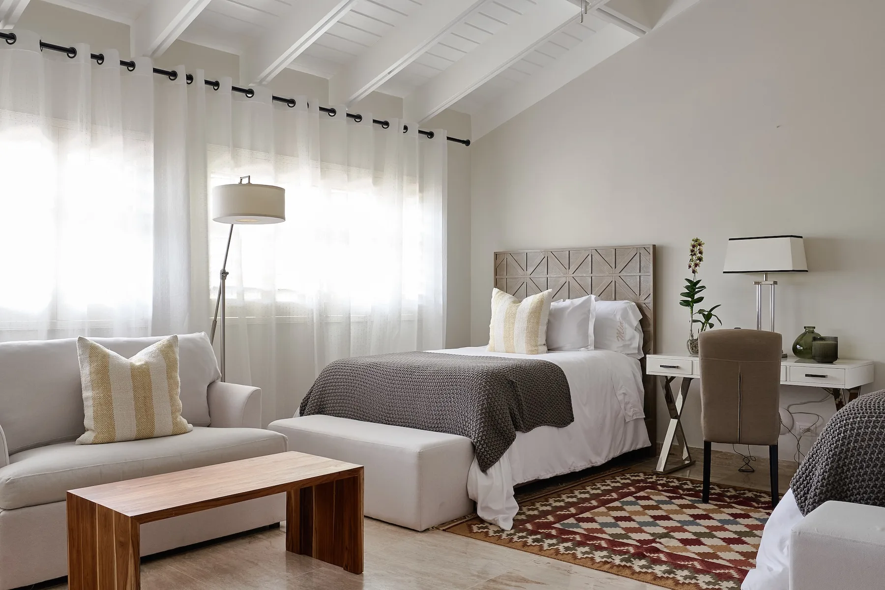 A bright bedroom with a bed, seating, and tall curtained windows.