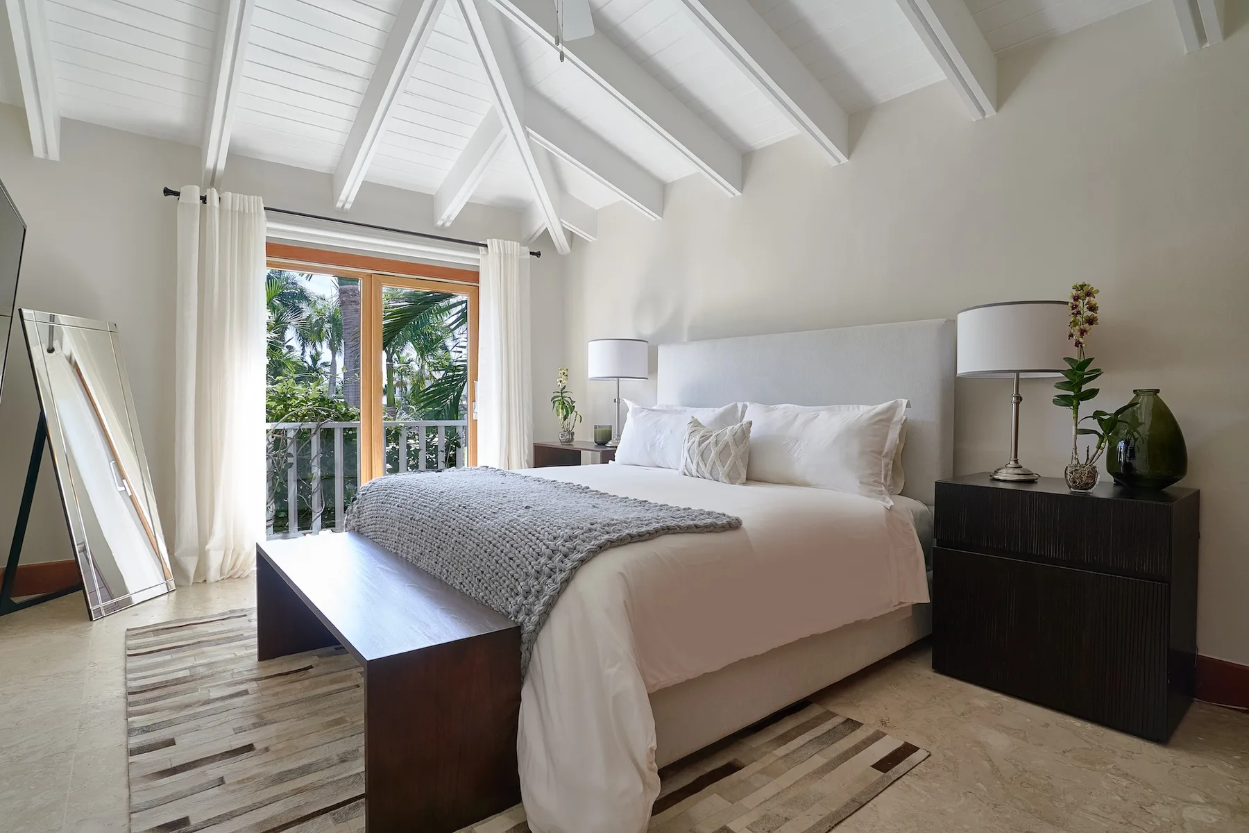 A bright bedroom with a large bed beneath a high, beamed ceiling.