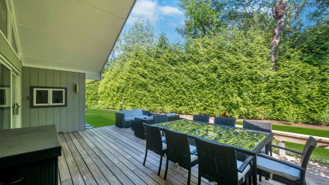 An outdoor deck has a dining table and seating beside a tall green hedge.