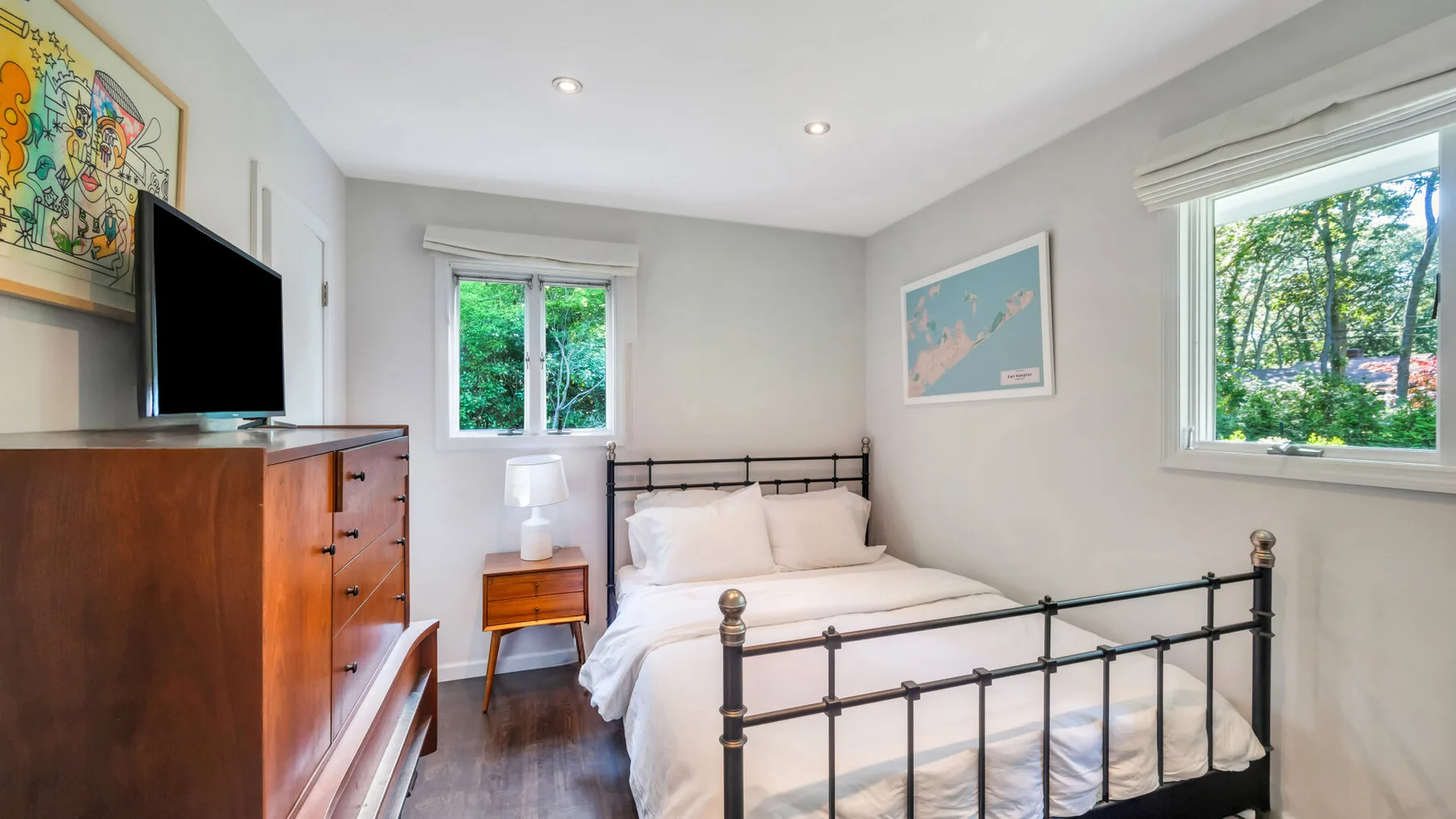 A bright bedroom with a metal bed, wooden dresser, and windows overlooking trees.