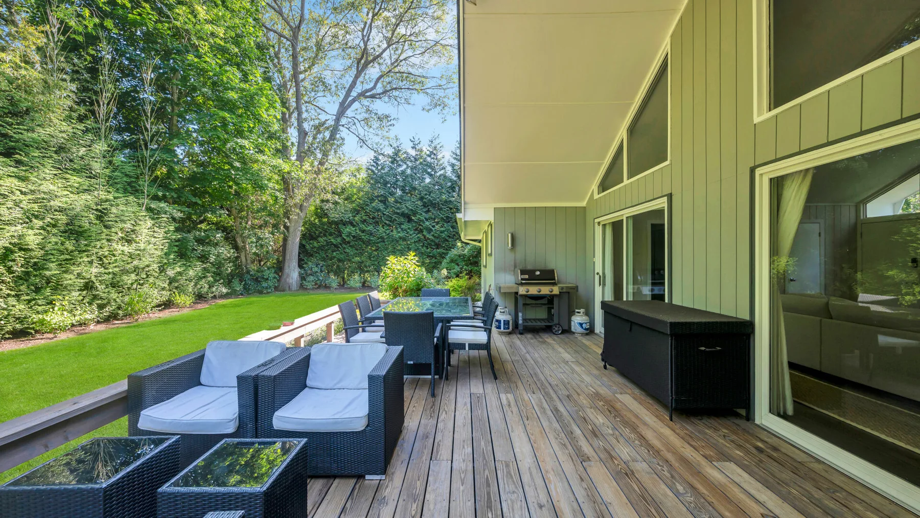 A covered outdoor deck with seating and a dining table overlooks a green lawn.