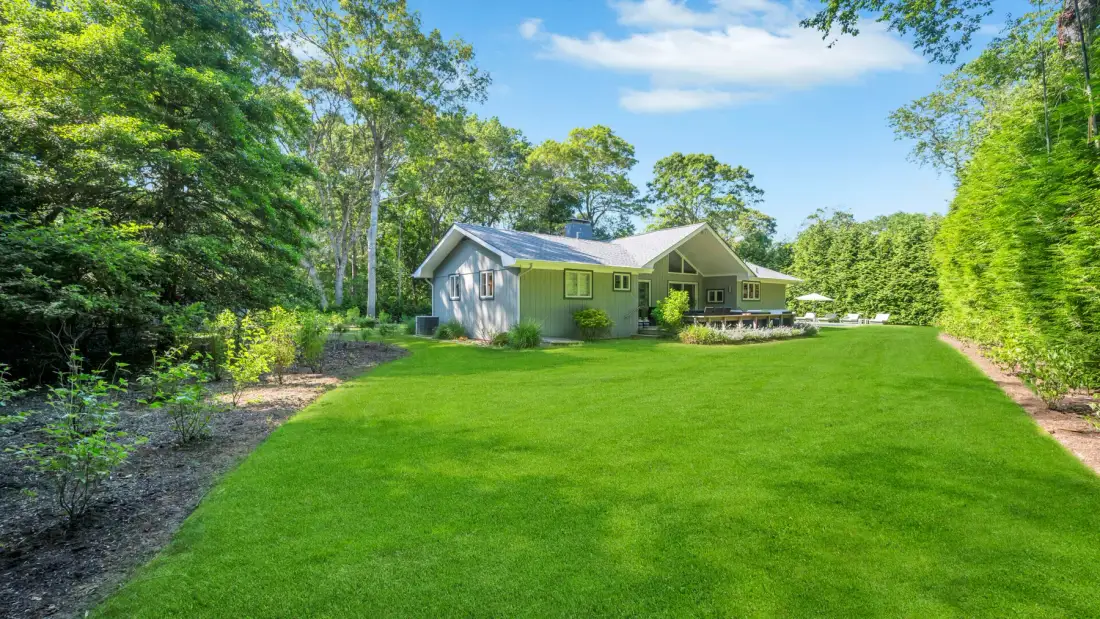 A house stands beyond a broad green lawn, surrounded by trees.