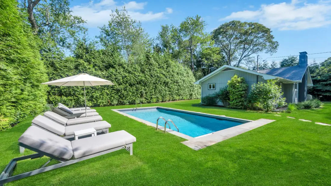 A backyard swimming pool beside a lawn and cushioned loungers under an umbrella.