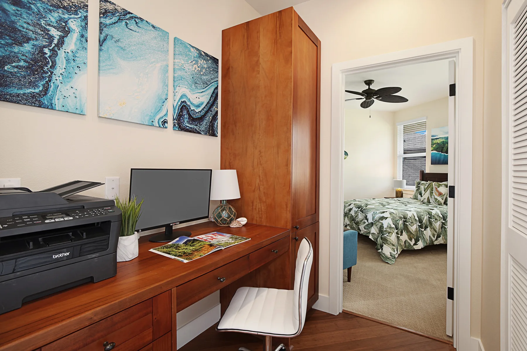 Un escritorio de madera con impresora y monitor está junto a la entrada abierta de un dormitorio.