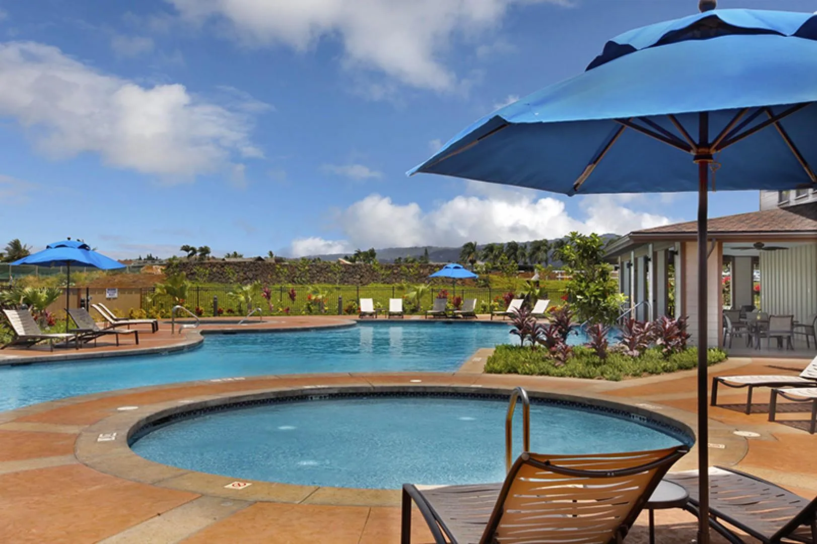 Una piscina exterior y otra circular más pequeña están junto a un patio ajardinado con sombrillas azules.