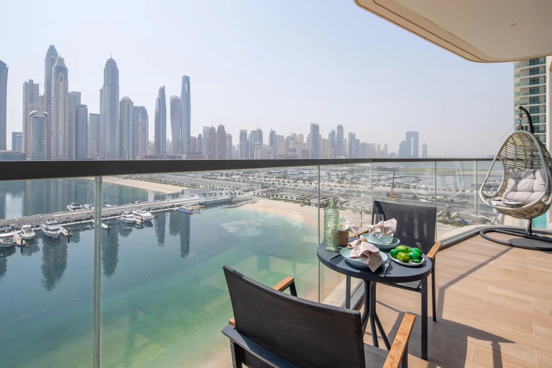 A waterfront balcony with outdoor seating overlooks a marina and distant high-rises.