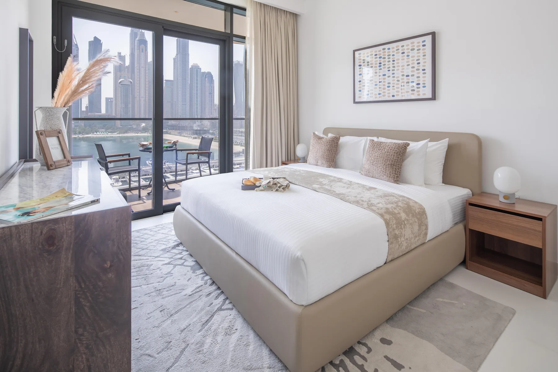 A bright bedroom has a large bed beside floor-to-ceiling windows overlooking city buildings.