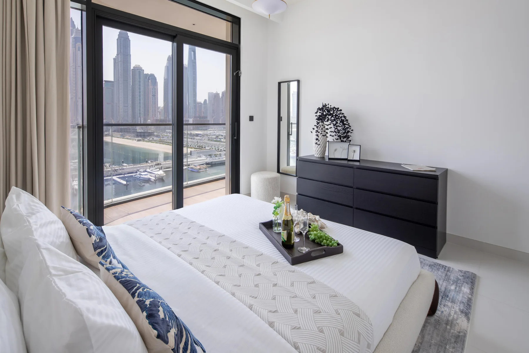 A bright bedroom with a bed beside large windows overlooking the city.