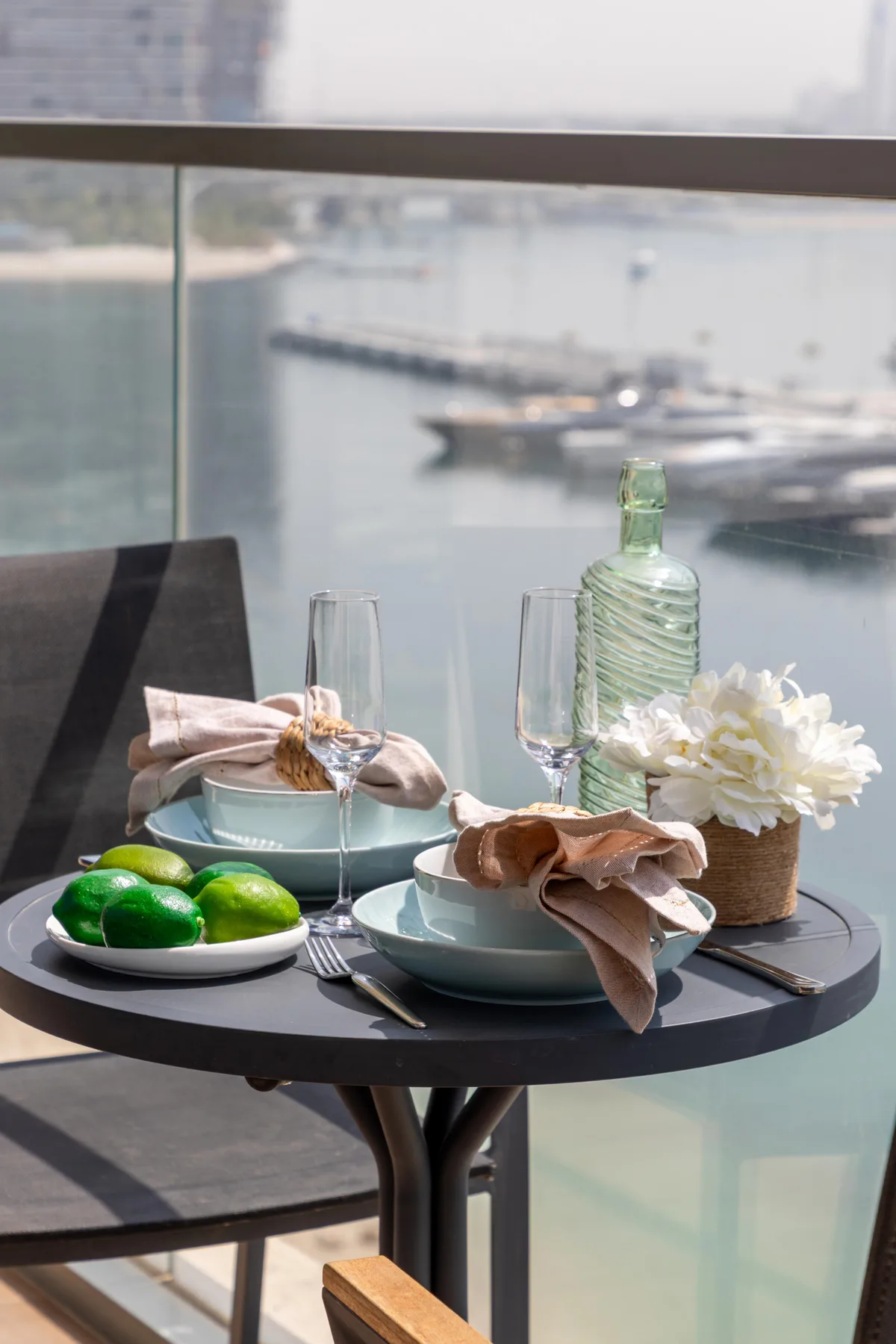 A small outdoor dining table is set with dishes, folded napkins, and flowers beside a glass railing.