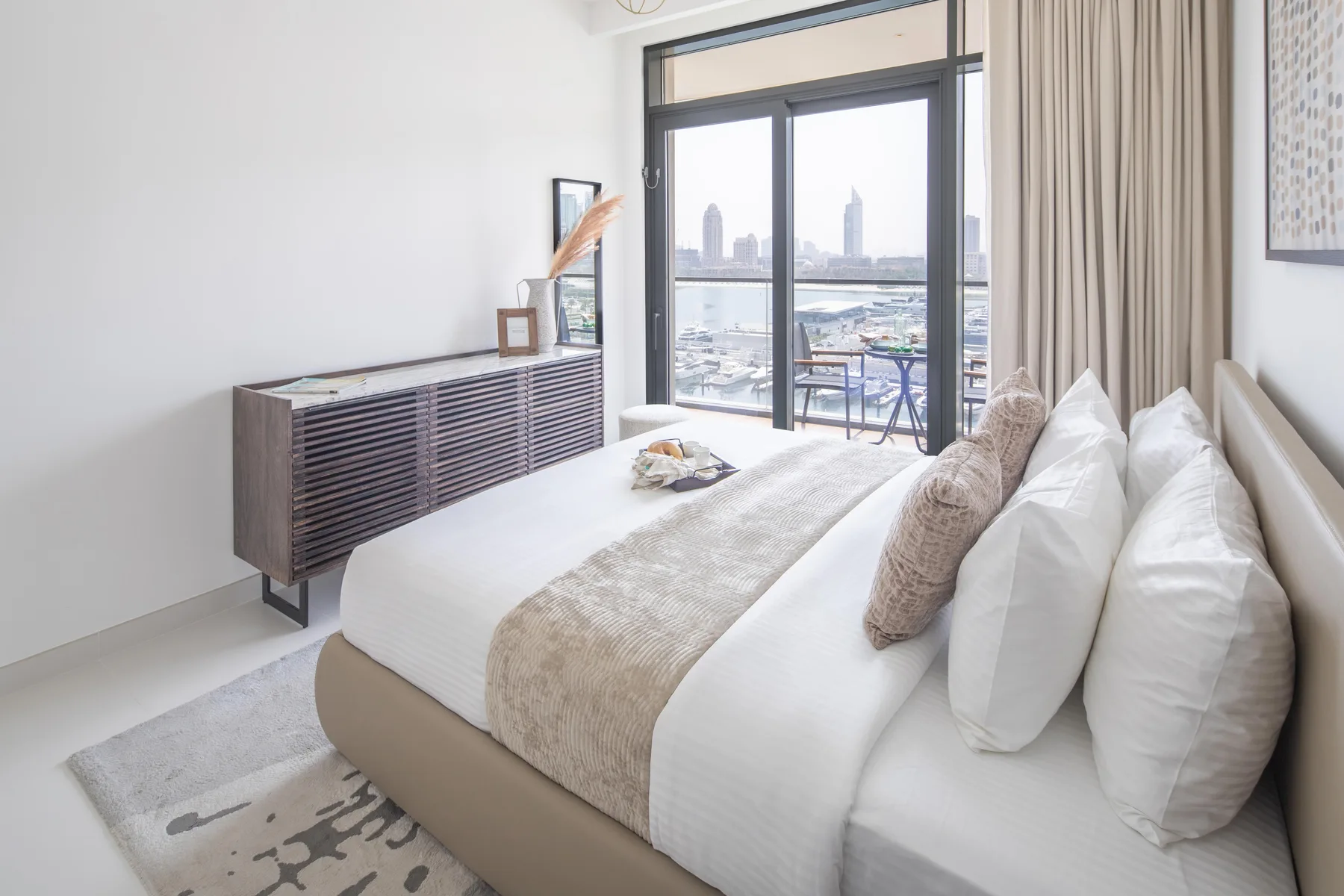 A bright bedroom with a neatly made bed beside large windows overlooking the city.