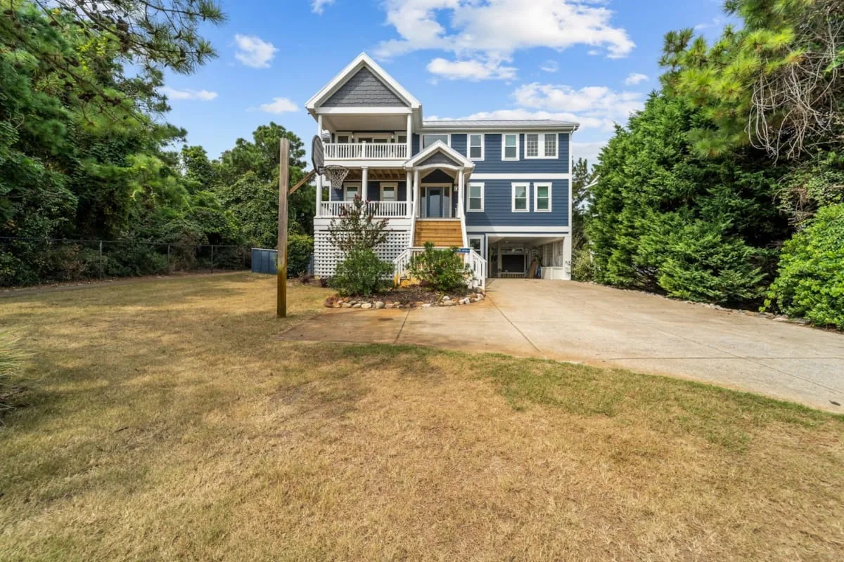 A large blue house stands beyond a broad driveway and dry lawn.