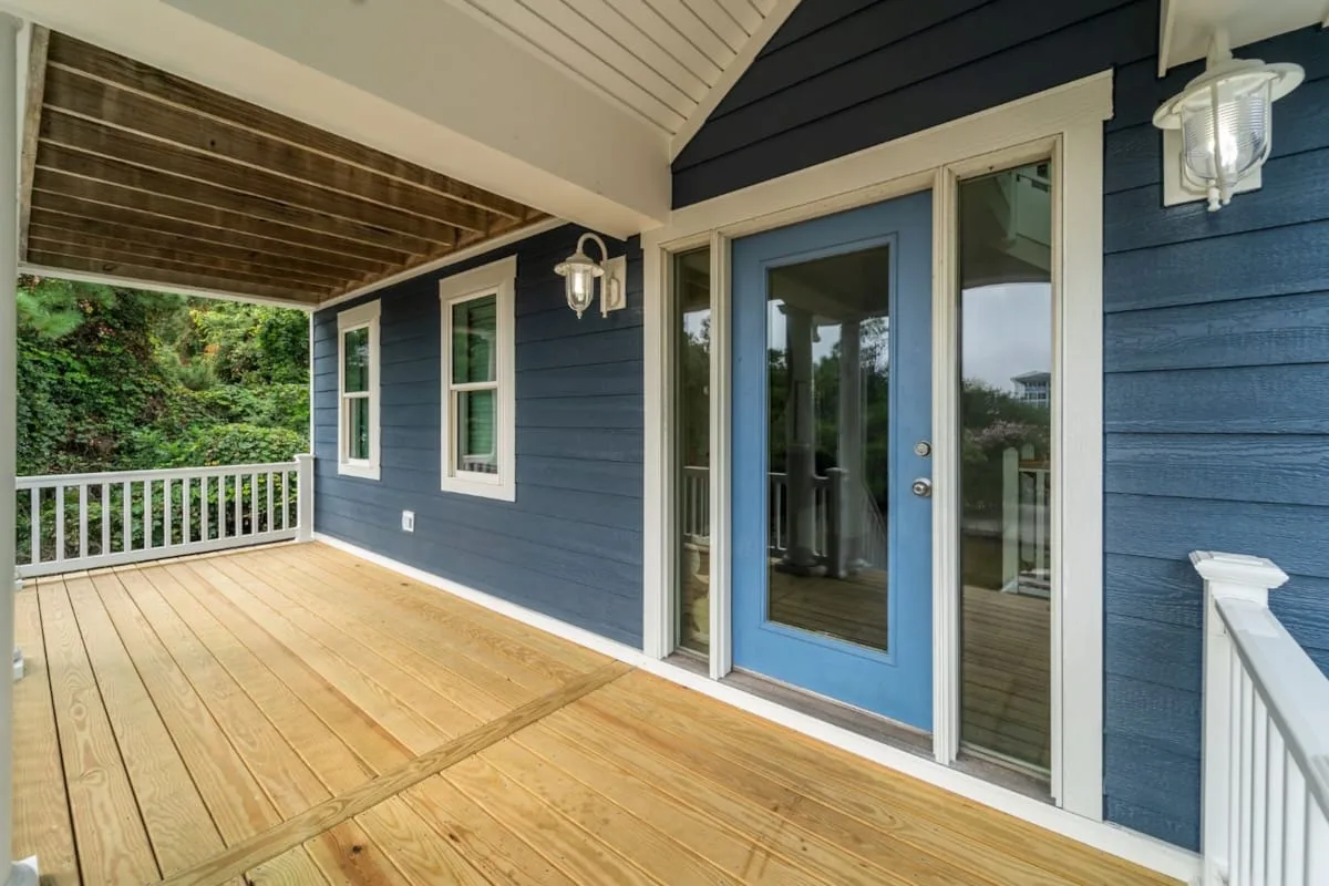 Covered wooden deck beside a blue house, with a glass-paneled door and white railings.