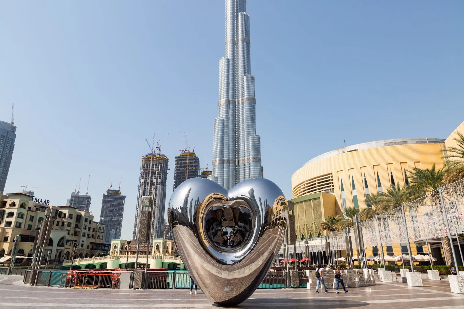 A heart-shaped reflective sculpture stands before a tall skyscraper in a city plaza.