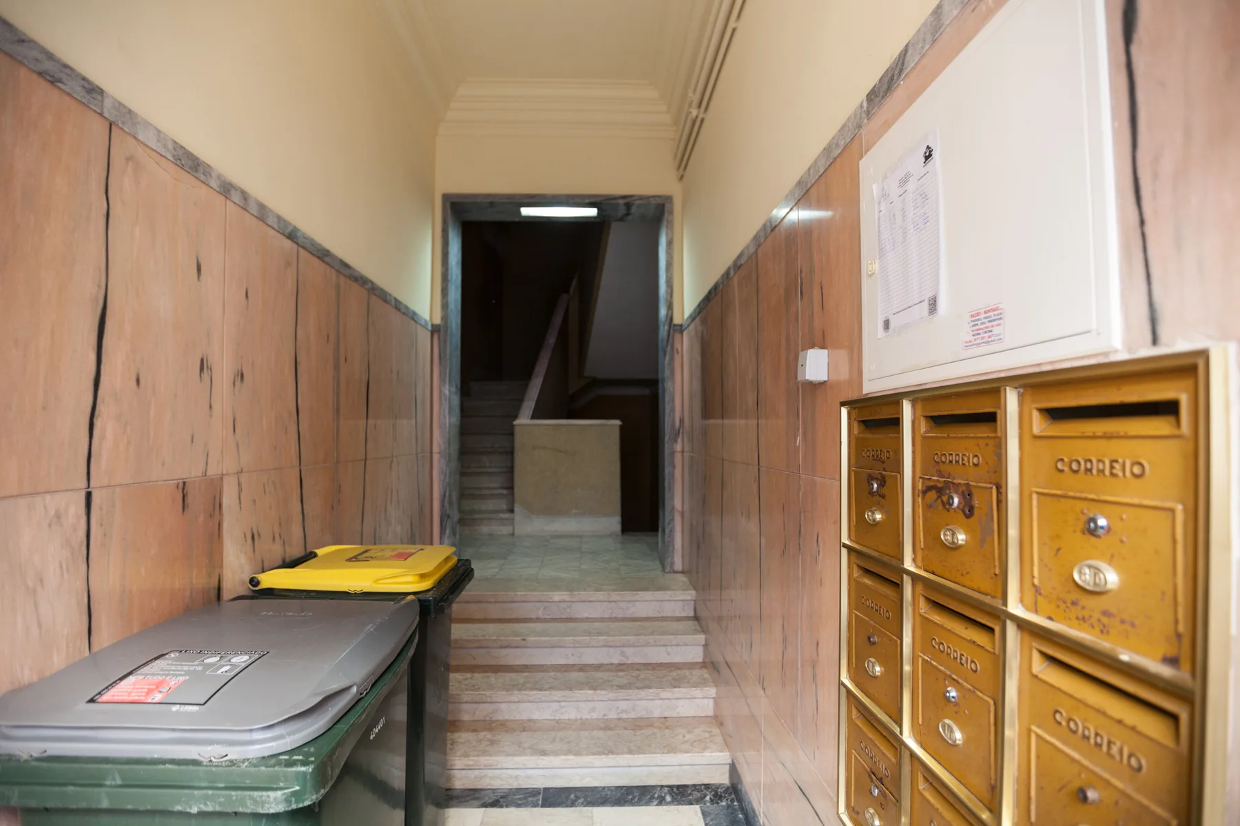 A tiled building entryway with mailboxes and waste bins beside a staircase.