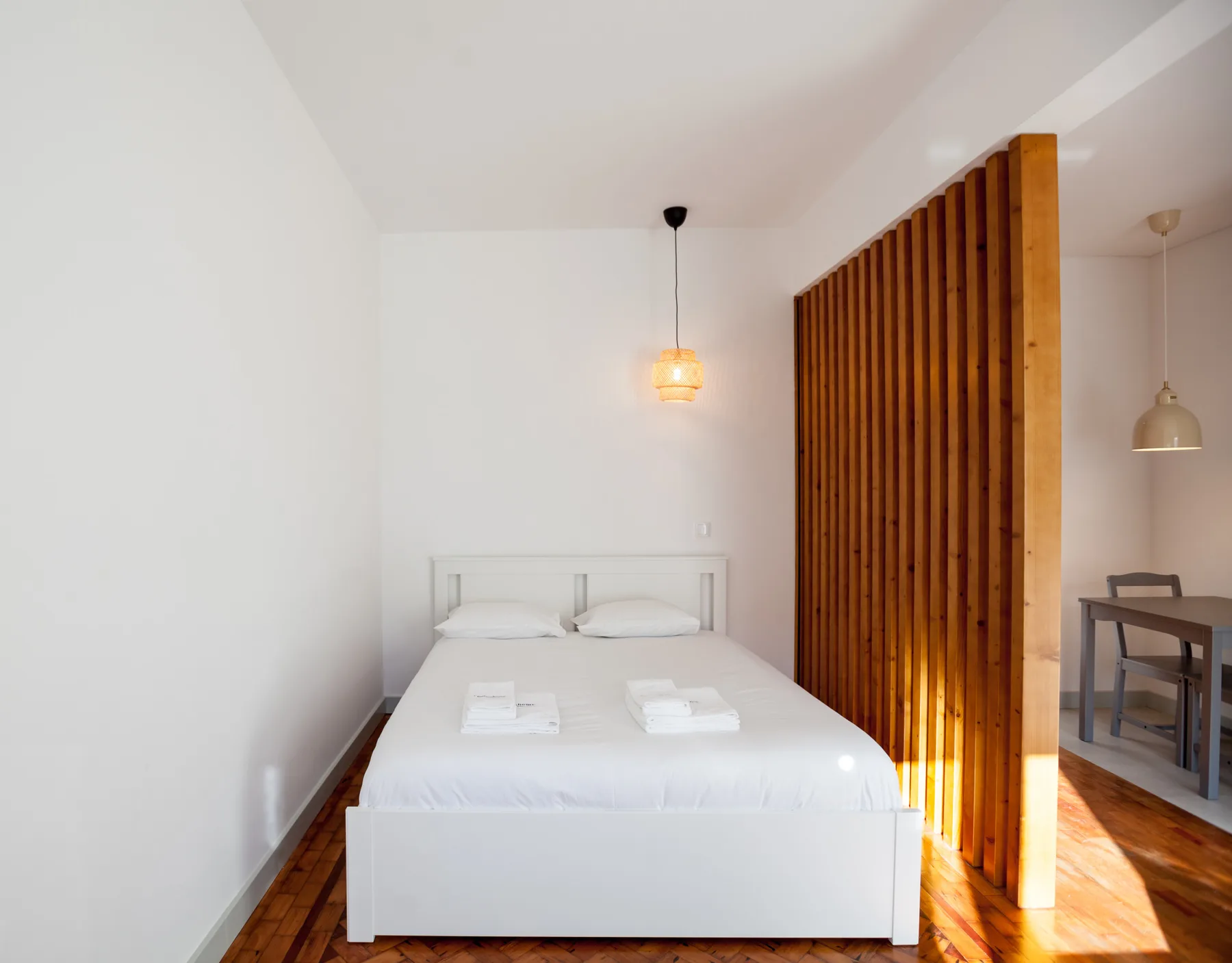 A bright studio bedroom with a white bed beside a wooden slatted divider.