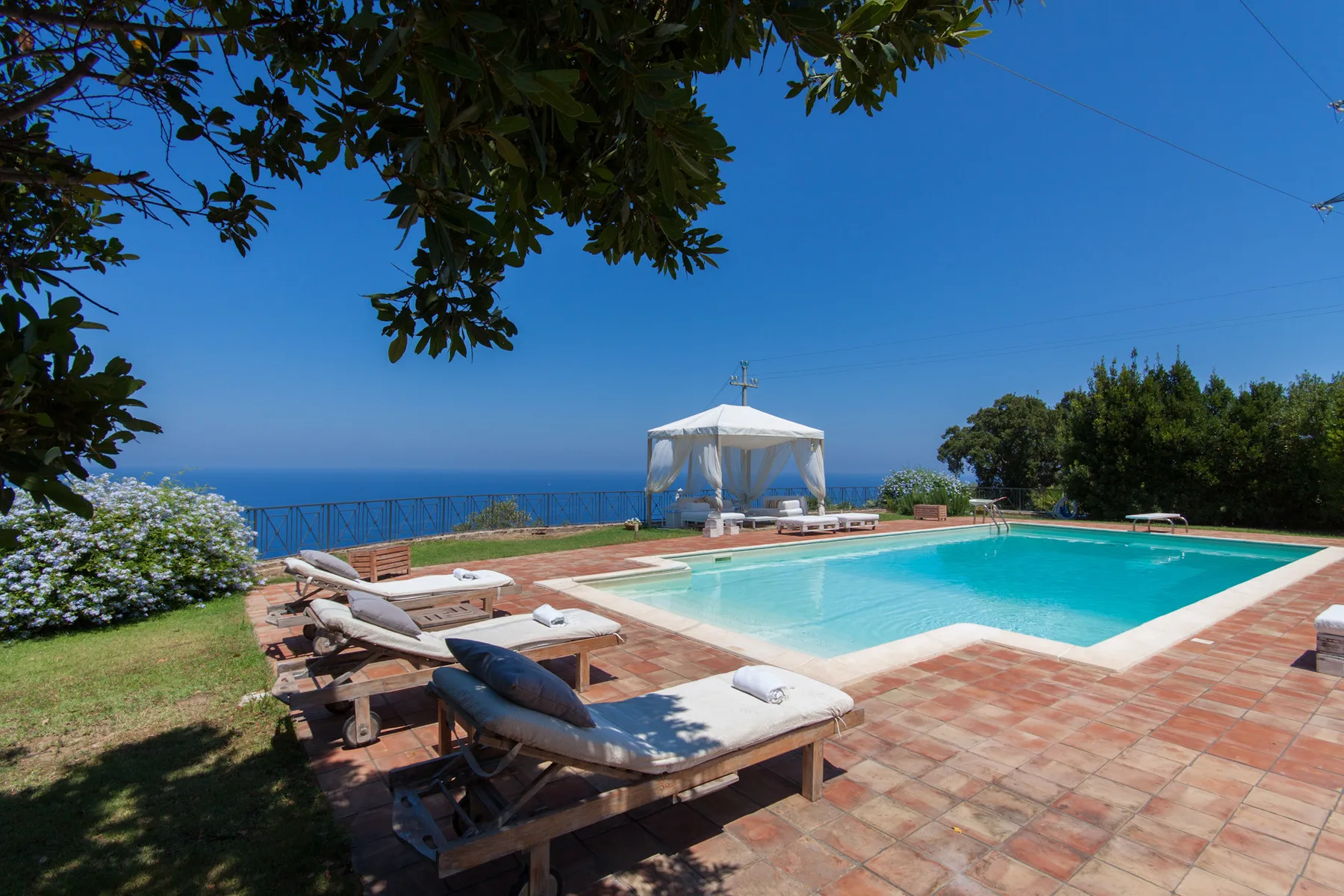 An outdoor swimming pool overlooks the sea, with lounge seating and a shaded canopy nearby.