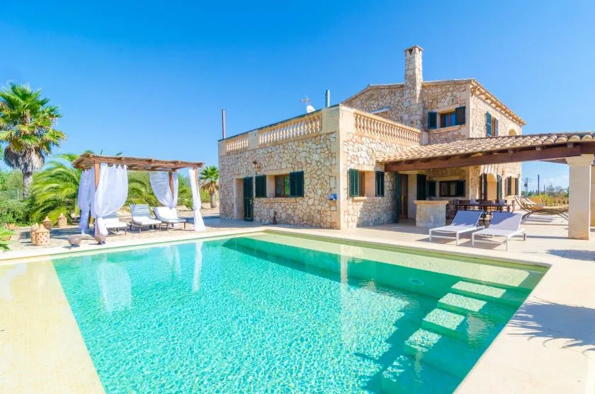 Una casa de piedra está junto a una piscina y una zona de estar a la sombra.