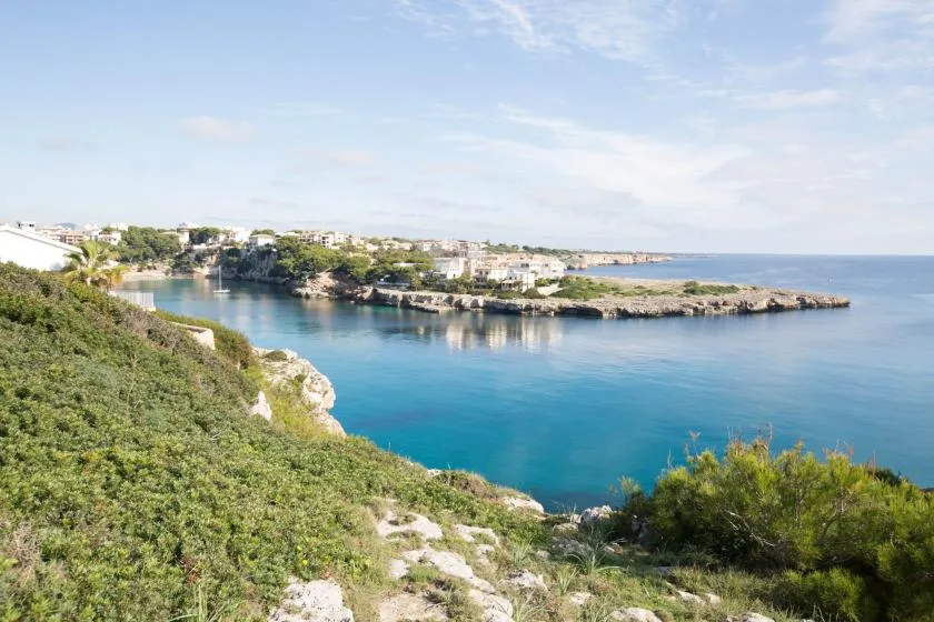 Bahía costera de aguas azules, orilla rocosa y edificios bajos junto a la costa.