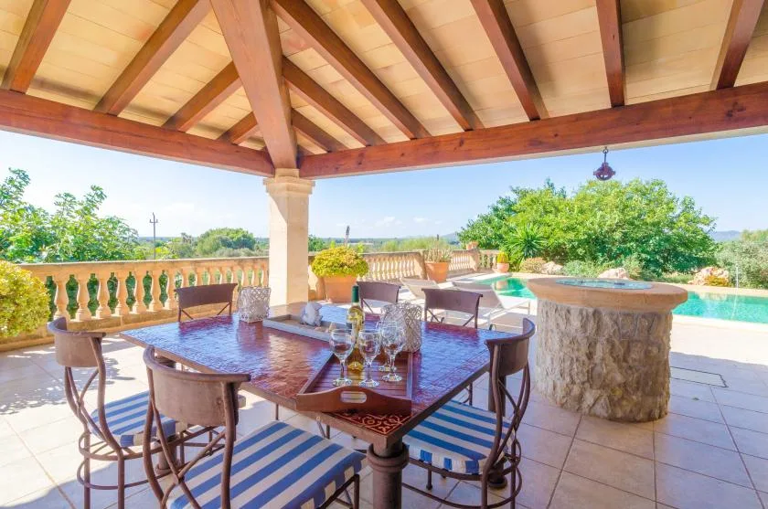 Una zona de comedor exterior cubierta con mesa y sillas da a una piscina y vegetación.