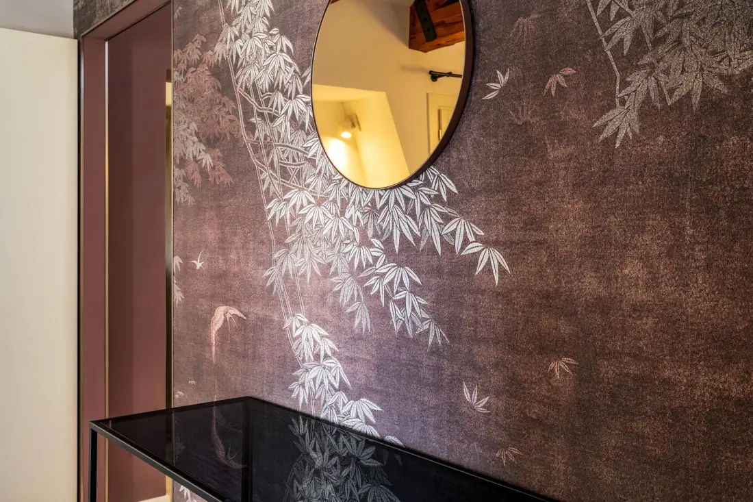 A hallway wall features botanical wallpaper, a round mirror, and a narrow console table.