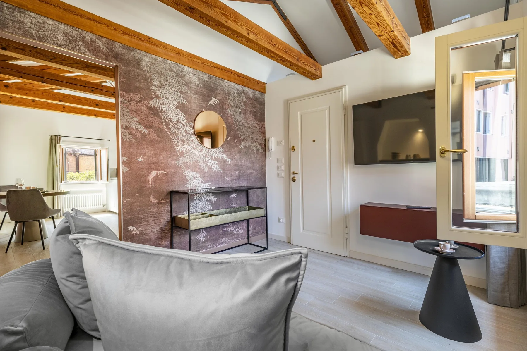 A bright living room with a gray seating area, wall-mounted television, and exposed ceiling beams.