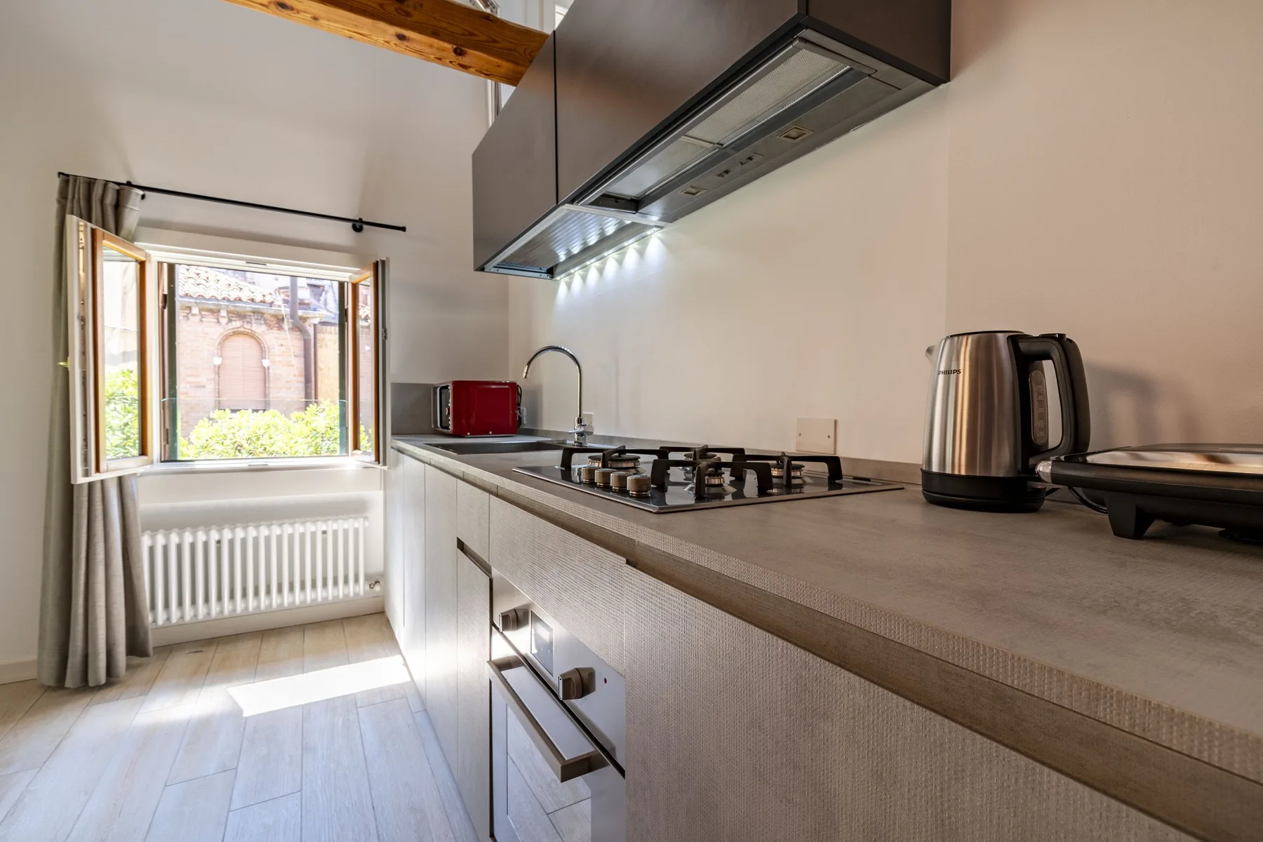 A kitchen with a countertop stove, sink, and kettle beside a window.