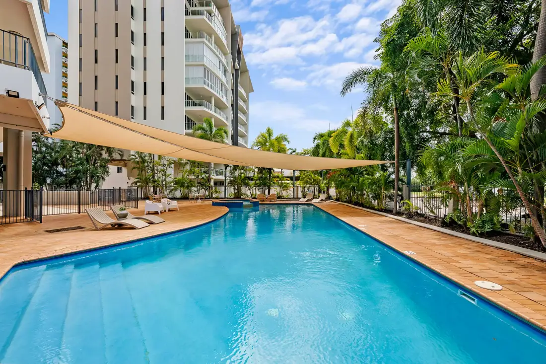 A swimming pool beside apartment buildings, with shaded seating and palm trees.