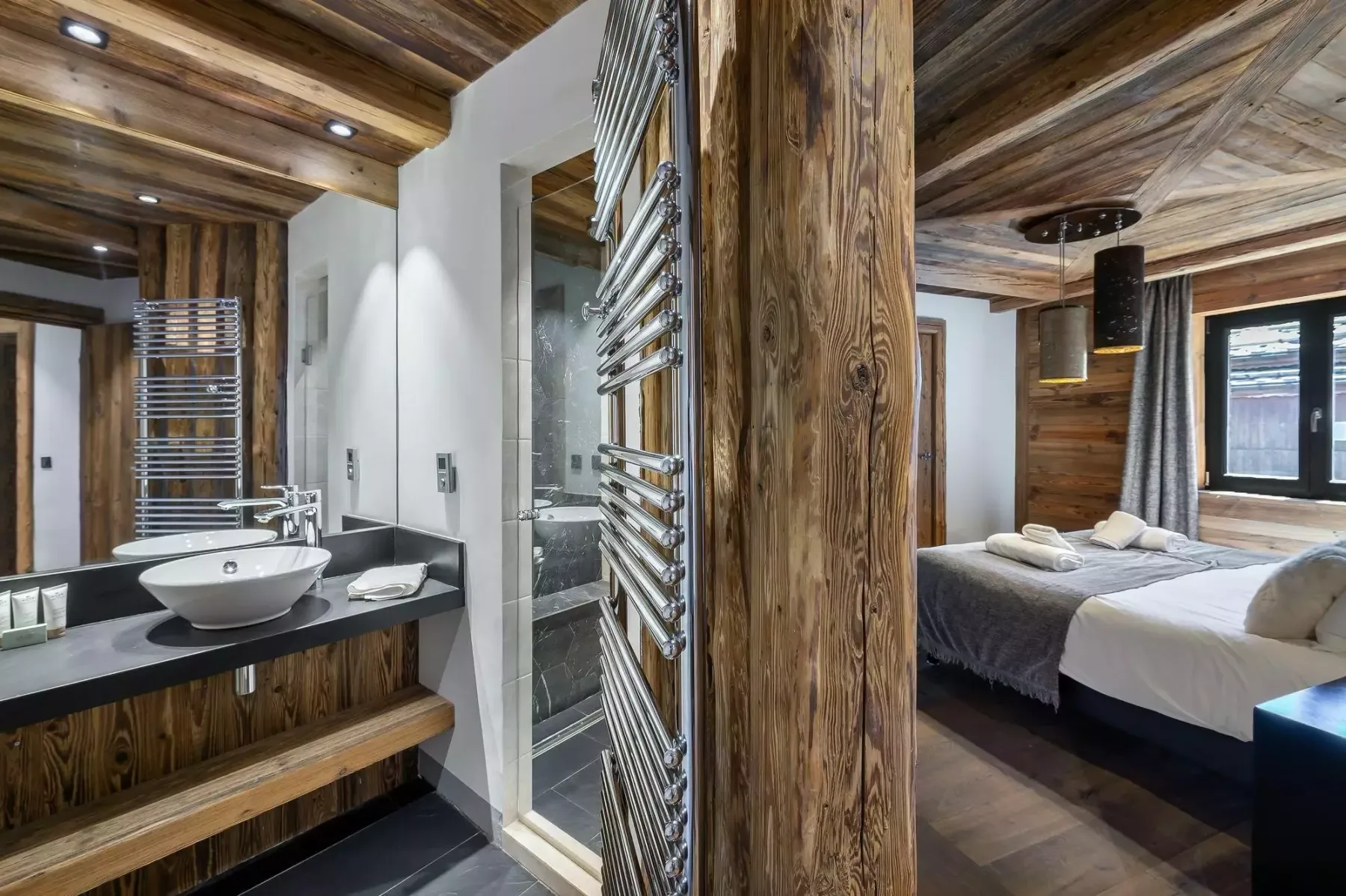 A bedroom and open bathroom feature wood finishes, a sink, and a bed beside a window.