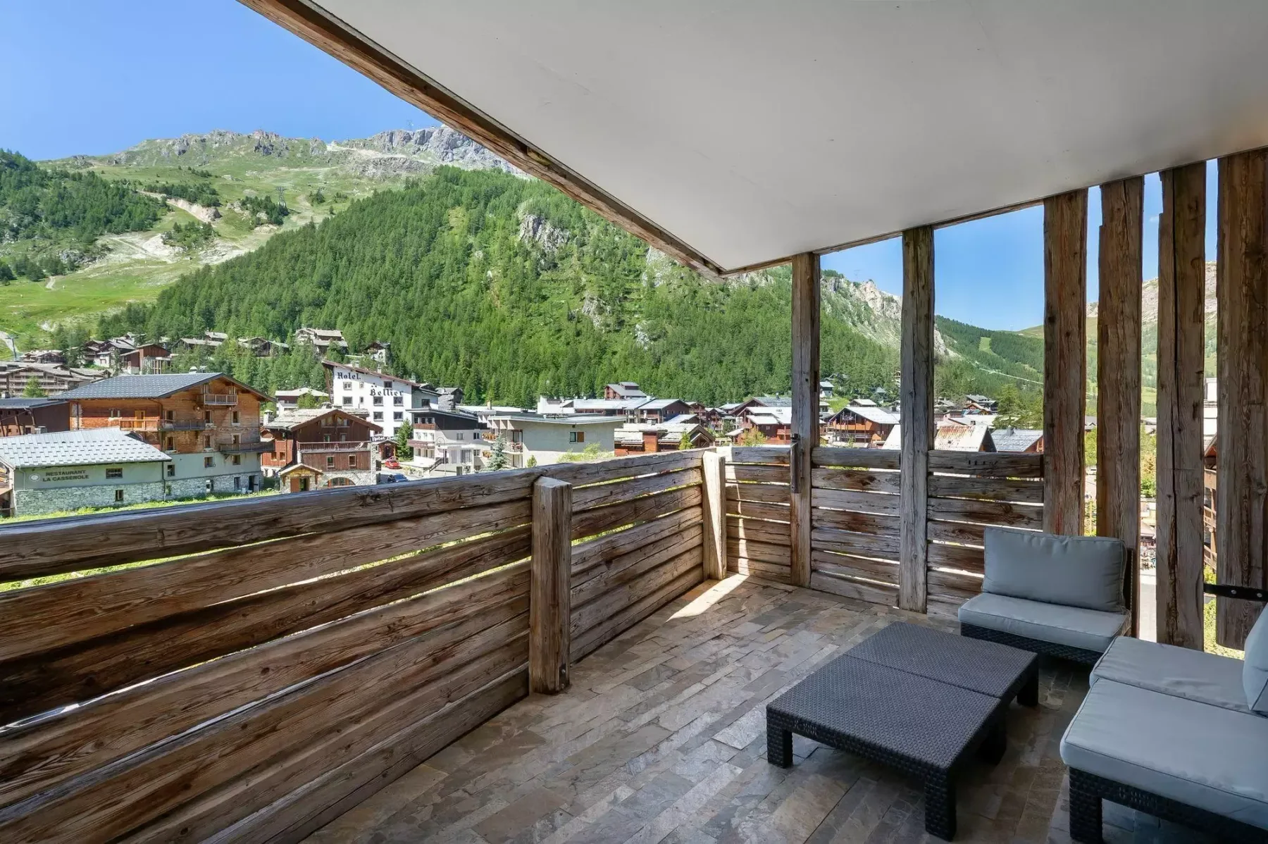 A wooden balcony with outdoor seating overlooks a mountain village and green hills.