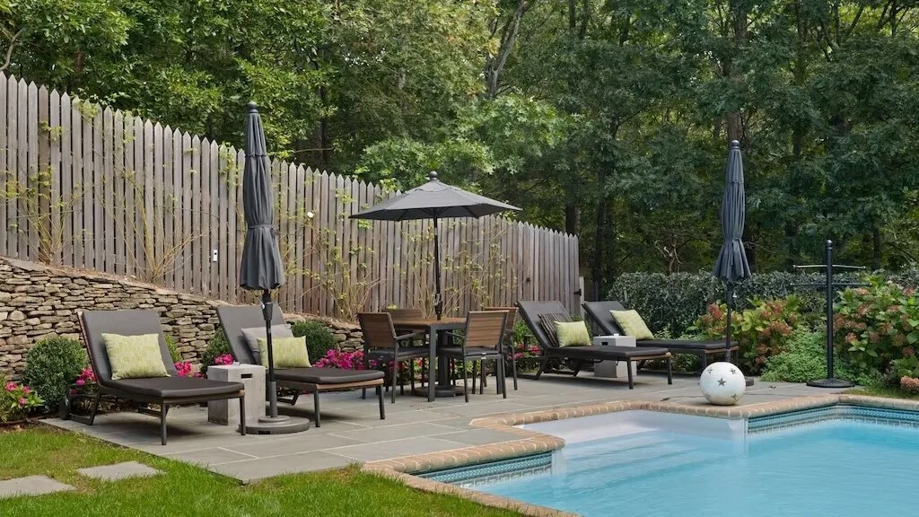 An outdoor pool area has patio seating and umbrellas beside a wooded fence.