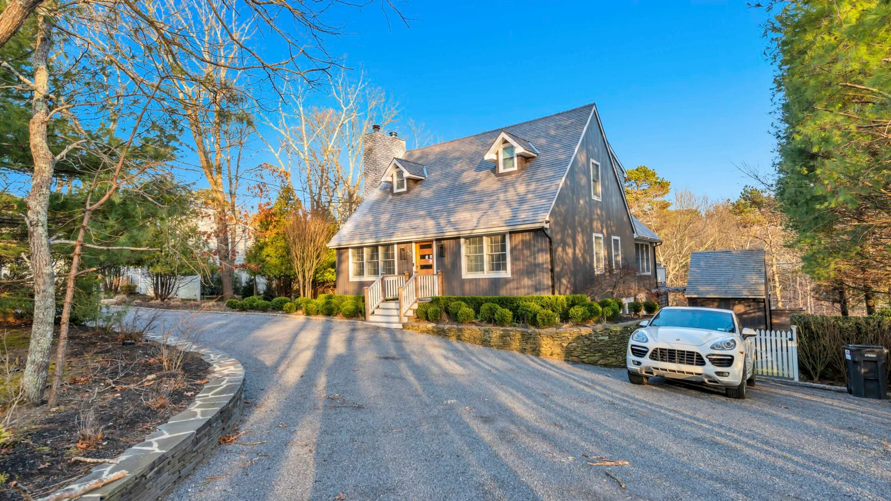 A dark-shingled house stands beside a curved driveway, with a car parked near the entrance.
