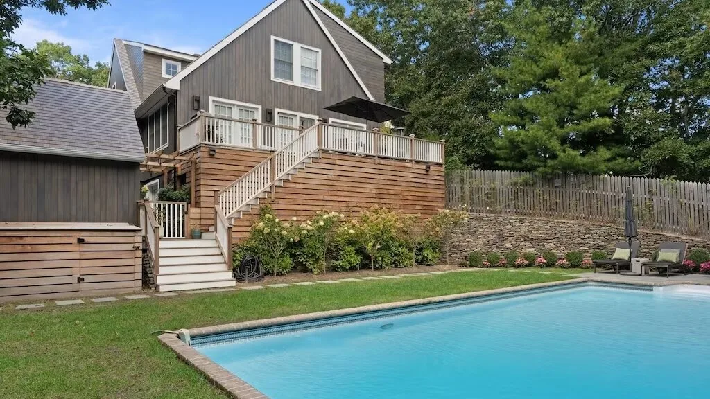 A backyard swimming pool sits beside a raised wooden deck and house.