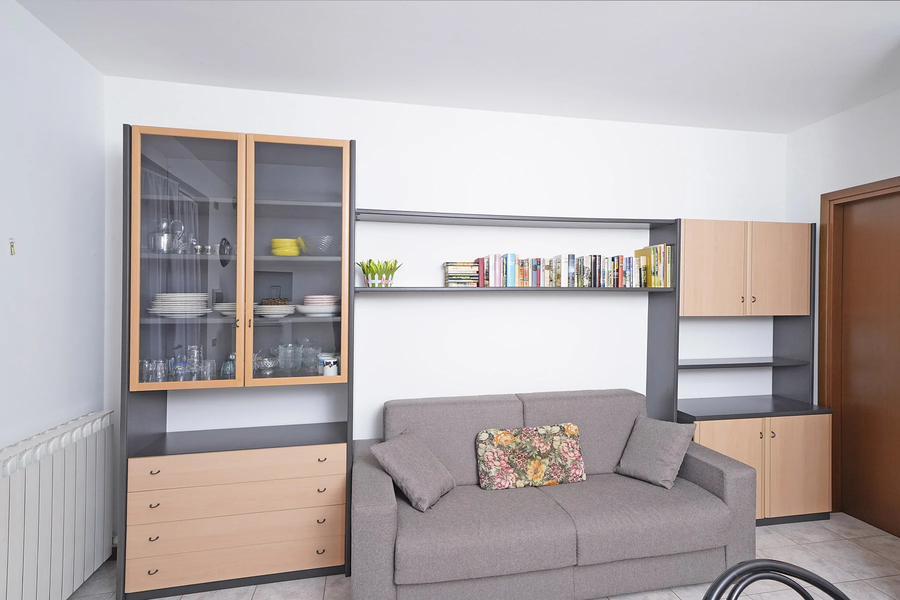 A living area with a gray sofa beneath shelves of books and cabinets.
