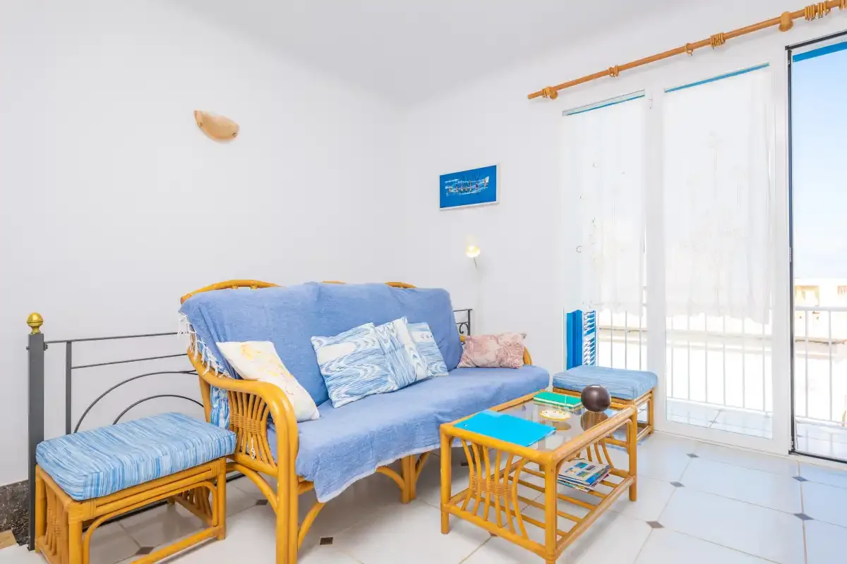 Una sala de estar luminosa con asientos azules, una mesa de centro de madera y ventanas grandes.