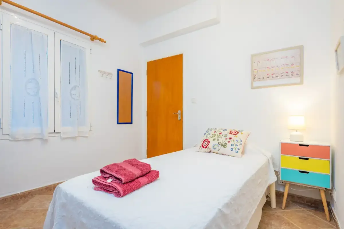 Un dormitorio luminoso con una cama, una mesita de colores y ventanas con contraventanas.