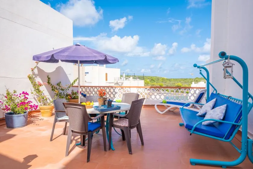 A sunny outdoor terrace with a dining table, purple umbrella, and hanging swing seat.