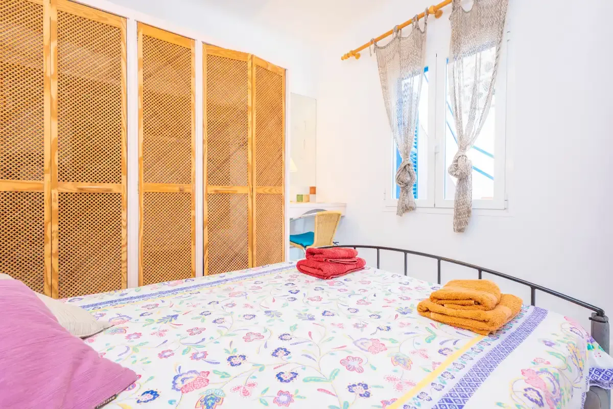 Un dormitorio con una colcha floral, puertas de armario tejidas y una ventana con cortinas.