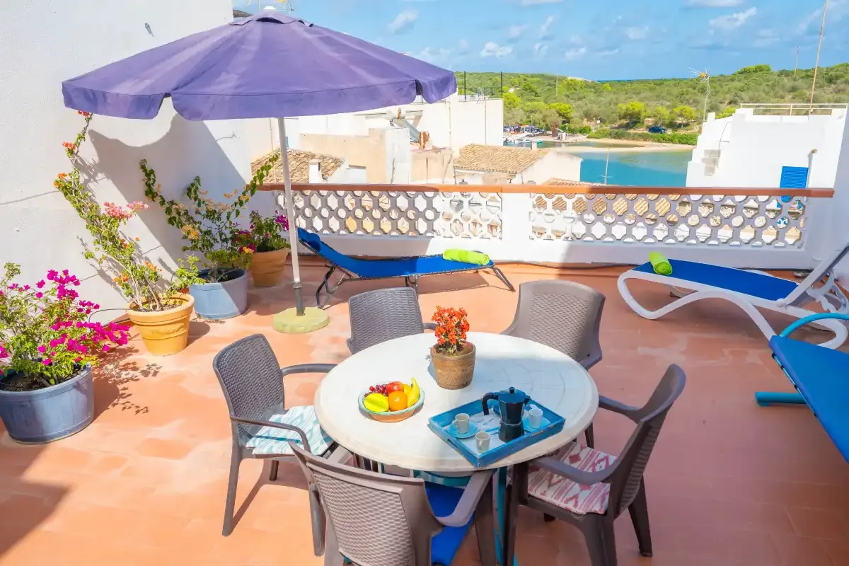 Una terraza soleada tiene una mesa redonda y asientos bajo una sombrilla morada.