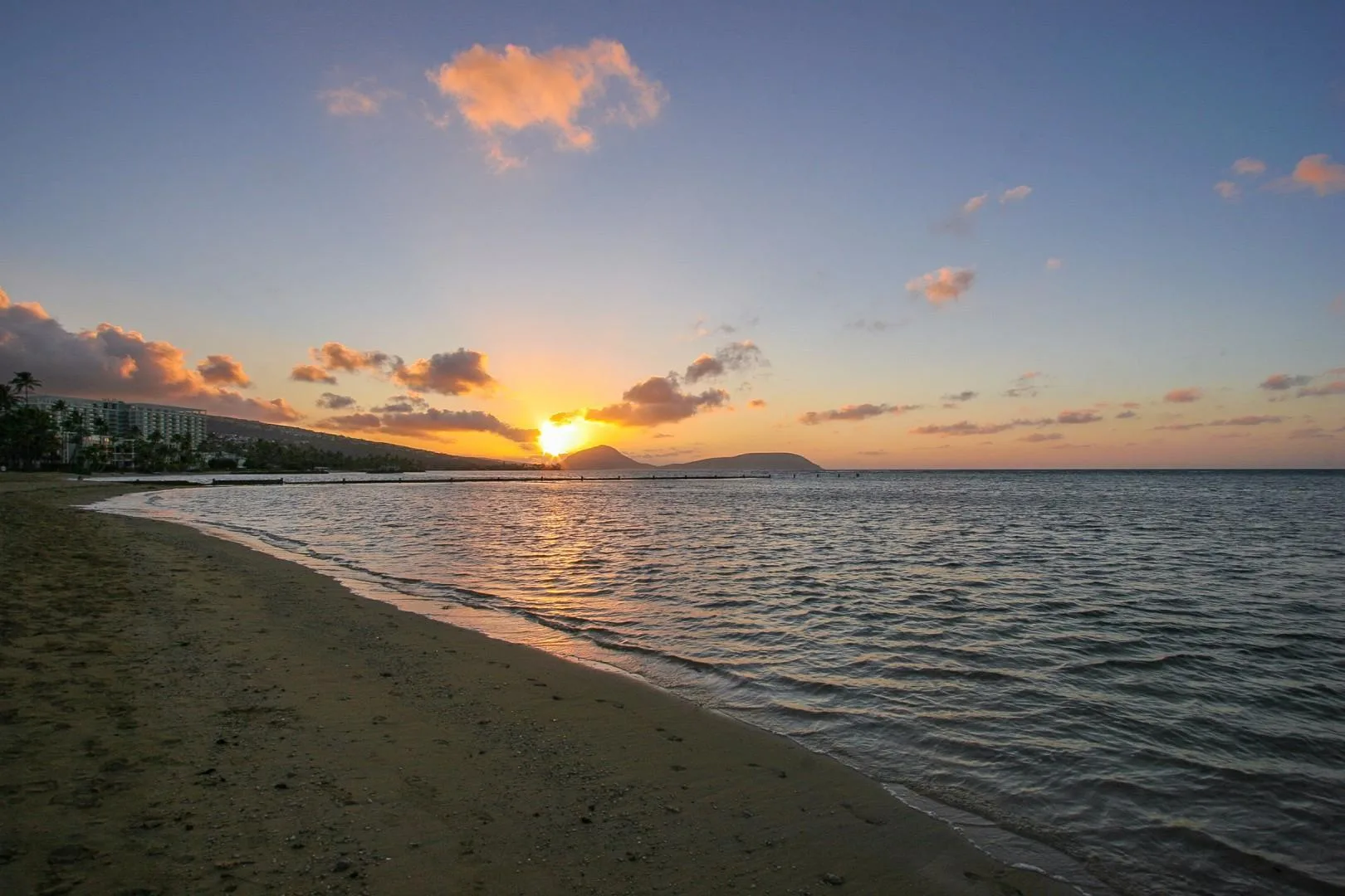 A sandy beach meets calm water beneath a colorful sunset sky.